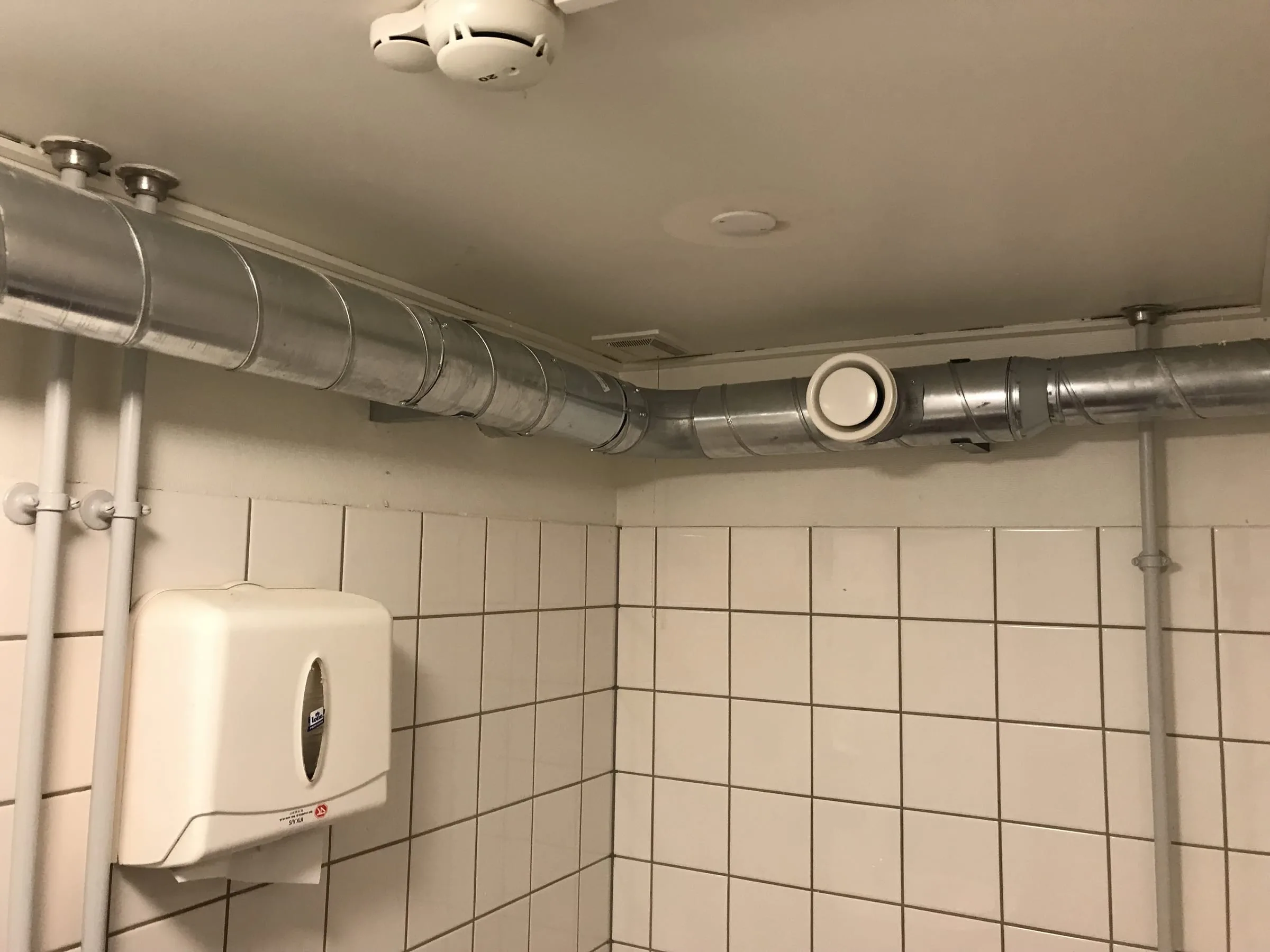 Rund udsugningskanal med en ventil langs loftet over hvide fliser på et toilet, Ravnsholtskolen, Allerød